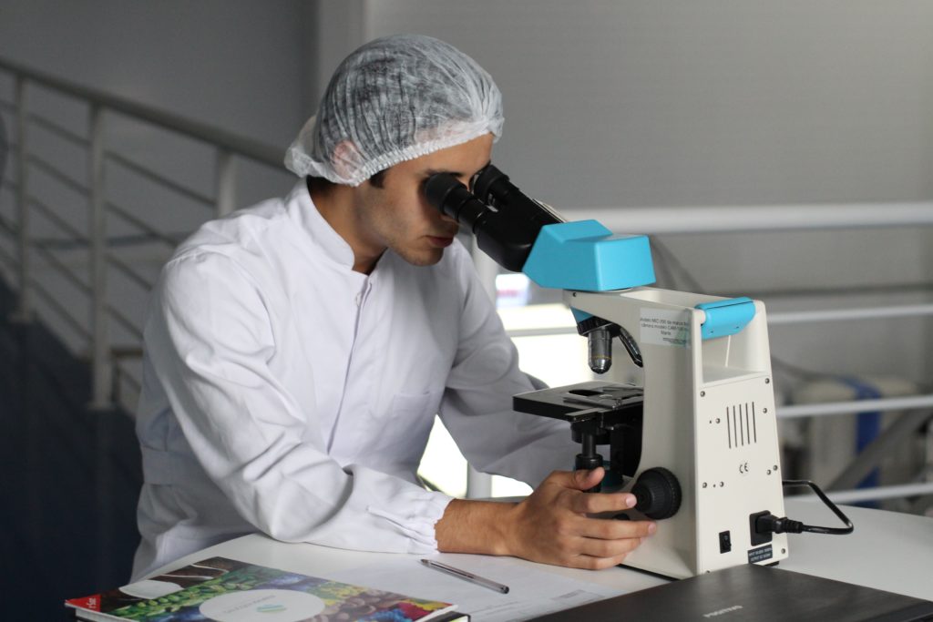 Scientist wearing white lab coat and net hat, looking down a microscope