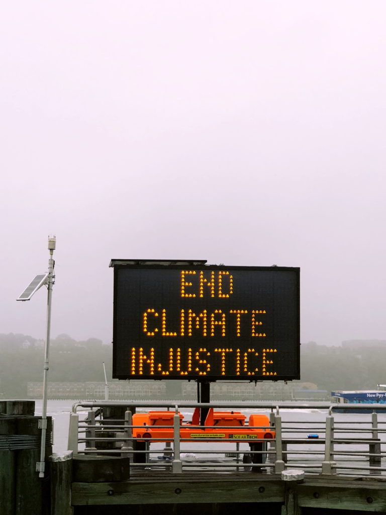 Electronic sign with the slogan "End Climate Injustice" sited by a habour on a cloudy day