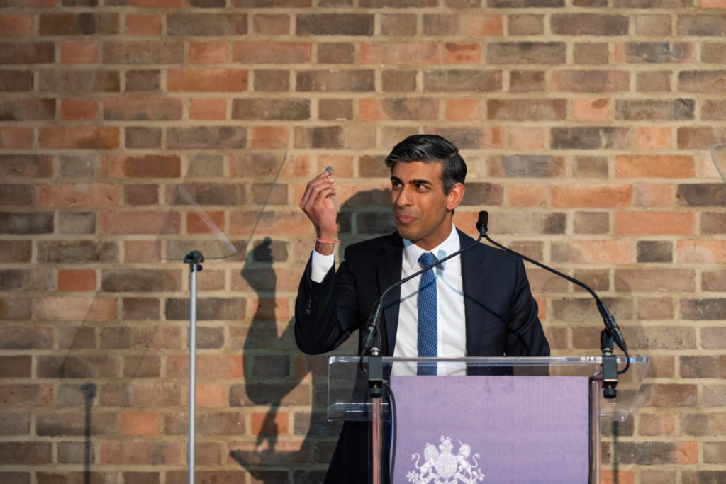 Rishi Sunak holding a £1 coin while making a speech